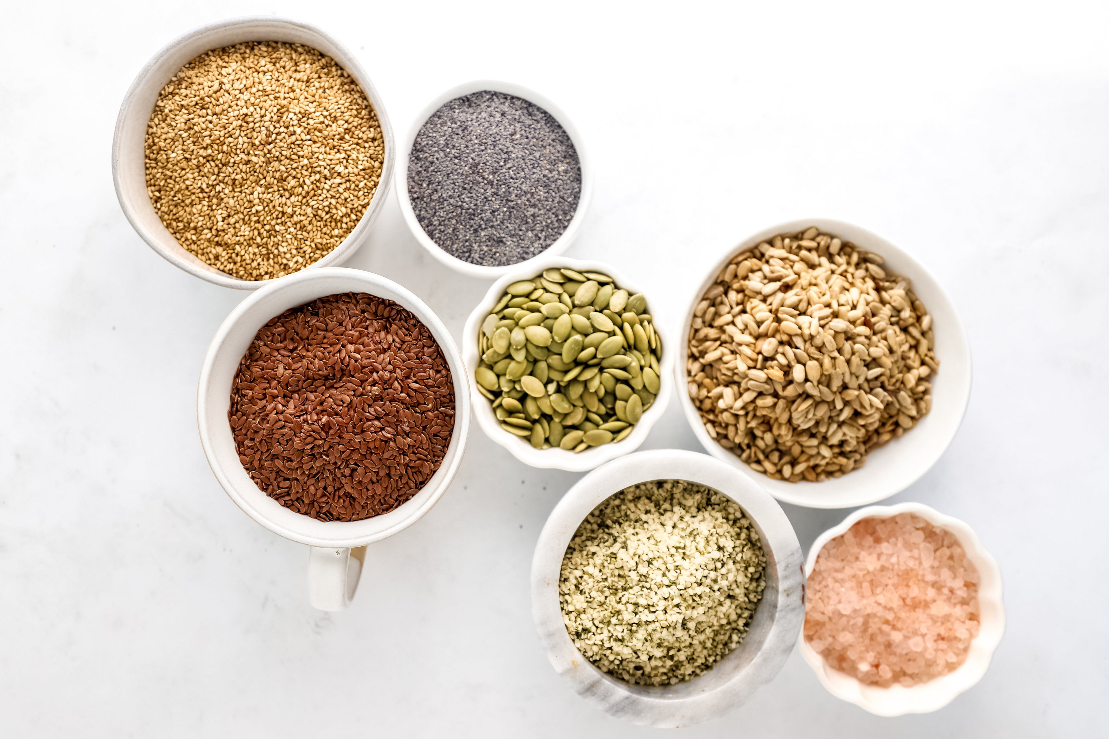 Bowls of sesame seeds, flaxseeds, hemp seeds, sunflower seeds, pumpkin seeds, poppy seeds, and Himalayan pink salt, showcasing the wholesome ingredients used in Ella’s Flats seed crackers.
