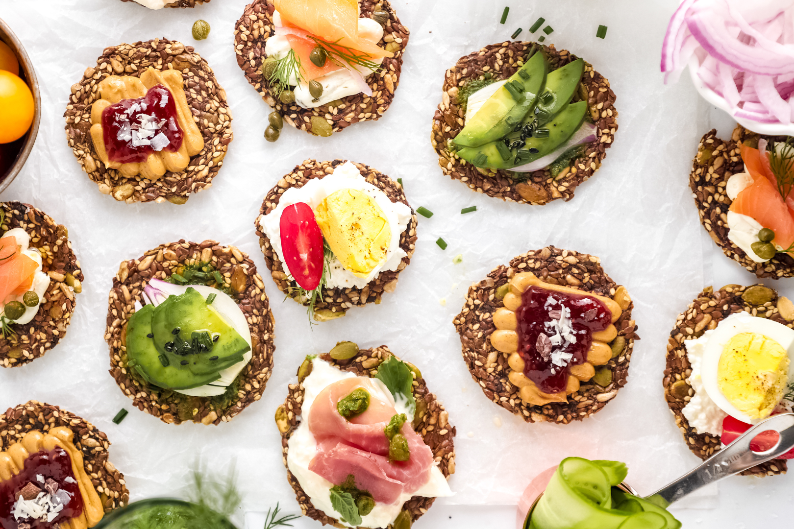 Ella’s Flats seed crackers topped with avocado, smoked salmon, eggs, peanut butter, jam, and fresh vegetables, showcasing versatile and healthy snack or meal ideas.