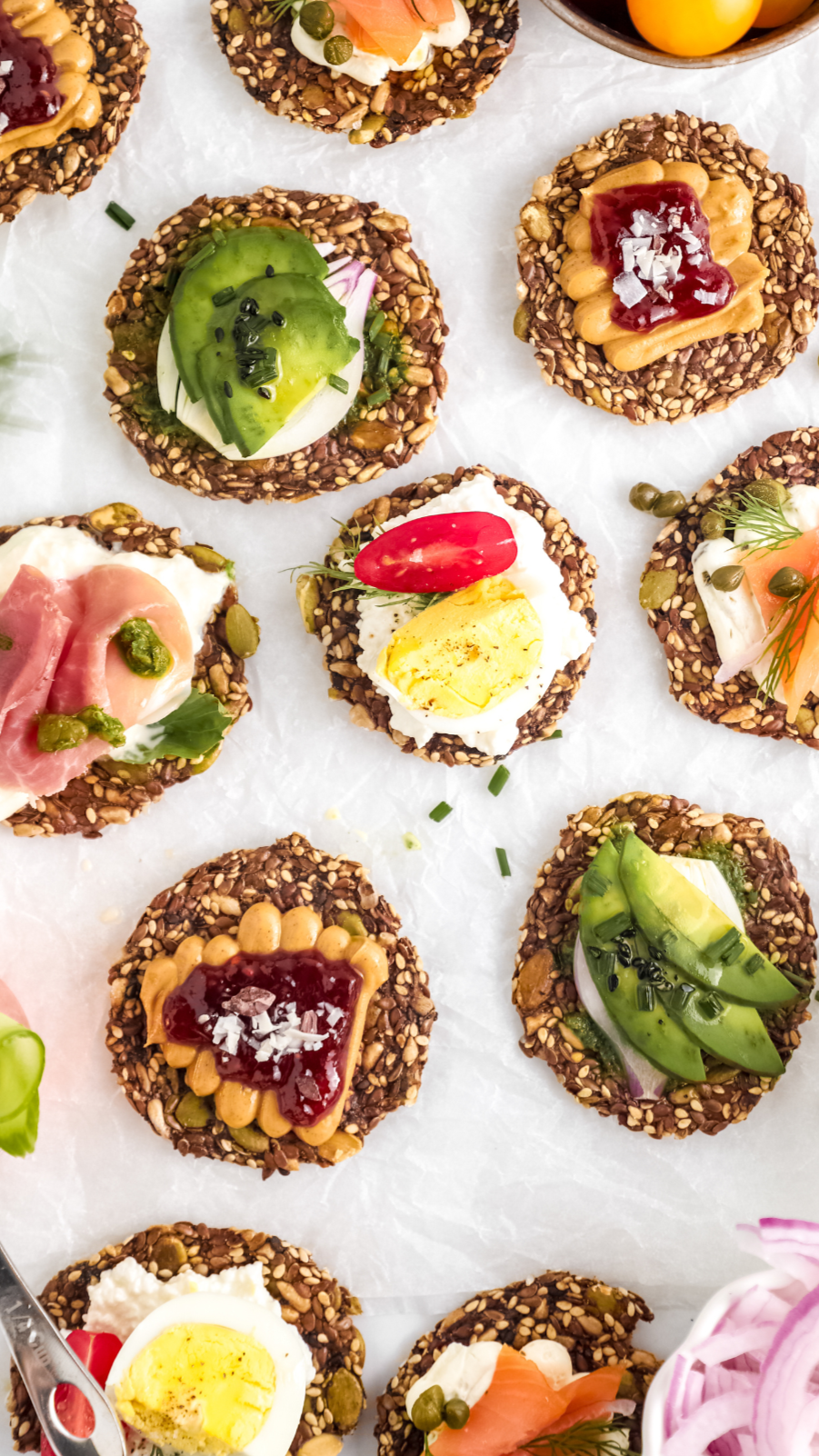 Ella’s Flats seed crackers topped with avocado, smoked salmon, eggs, peanut butter, jam, and fresh vegetables, showcasing versatile and healthy snack or meal ideas.